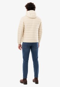 Giacca imbottita trapuntata beige chiaro con cappuccio. Presenta cuciture orizzontali e orlo elastico. Indossata sopra jeans blu e stivali marroni.