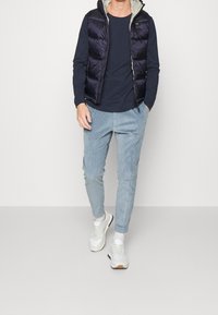 Mann, der ein navyblaues Langarmhemd, eine schwarze Steppweste, hellblaue Cordhosen, weiße Sneaker und weiße Socken trägt.