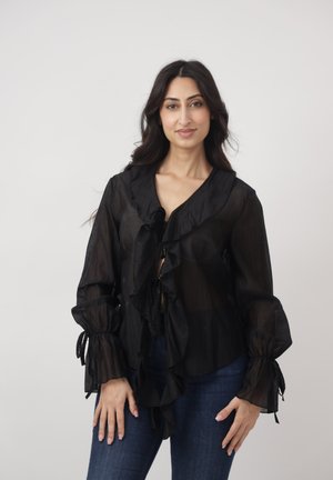 Kobieta o długich ciemnych włosach, ubrana w przezroczystą czarną bluzkę z falbankami i niebieskie dżinsy, stojąca na tle jednolitego jasnego tła.