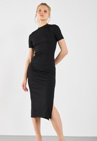 Crna rebrasta midi haljina s kratkim rukavima, prisna konstrukcija, bočni prorez i visoki izrez. Glatka tekstura i minimalistički stil.
