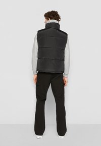 Sort puffer vest med høj krave, der har quiltede sektioner. Båret over en grå langærmet skjorte og kombineret med sorte bukser og hvide sneakers.