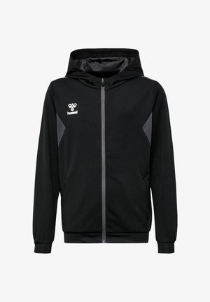 Jugendlicher steht mit den Händen in der Hüfte, trägt schwarzen Reißverschluss-Hoodie, passende Shorts, weiße Socken und schwarze Turnschuhe vor einfachem Hintergrund.