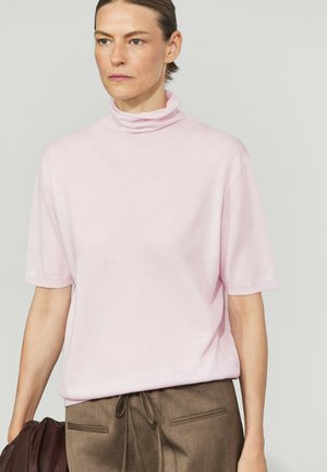 Femme aux cheveux courts portant un pull à col roulé manches courtes rose clair et un pantalon marron, tenant une veste marron dans une main.