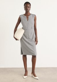 Robe ajustée à carreaux vichy noir et blanc avec un design sans manches, un décolleté en V et une taille ceinturée. Associée à un sac à main crème et des talons blancs.