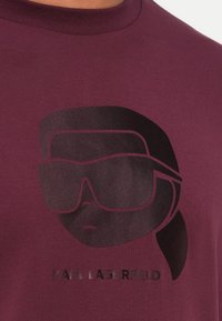 Stilisert svart grafikk av en person med hestehale som har på seg store solbriller på en bordeaux skjorte, med teksten "KARL LAGERFELD" under.