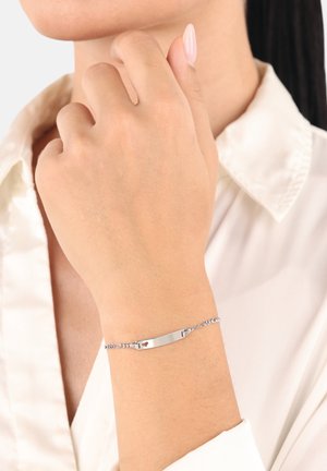 Bracciale d'argento con una barra piatta e un piccolo accento a forma di cuore, caratterizzato da un design a catena. Indossato su un polso contro una camicetta di colore chiaro.