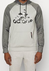 Sweat à capuche gris clair avec manches raglan gris foncé, arborant un grand logo Adidas noir réfléchissant et une poche kangourou à l'avant avec fermeture éclair.