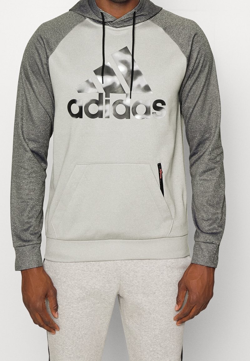 Sweat à capuche gris clair avec manches raglan gris foncé, arborant un grand logo Adidas noir réfléchissant et une poche kangourou à l'avant avec fermeture éclair.