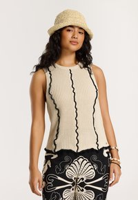 Beige ribots trikotāžas topiņš ar melnu volānu apdari; saskaņots ar melnām rakstainām platām biksēm un salmiņu cepuri.