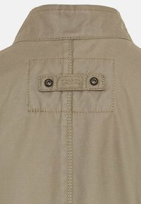 Beige jakke krave i en struktureret stof. Har et rektangulært mærke med "camel active" branding og to metaltrykknapper.