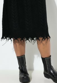 Schwarzes Strickkleid mit einem strukturierten Zopfdesign und fransigem Saum. Kombiniert mit knielangen schwarzen Lederstiefeln mit Nietenakzenten.