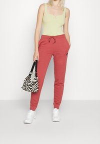 Kvinna iklädd en ljusgrön linne och röda Nike joggingsbyxor, håller en väska med zebramönster och har vita sneakers på sig, står framför en vit bakgrund.