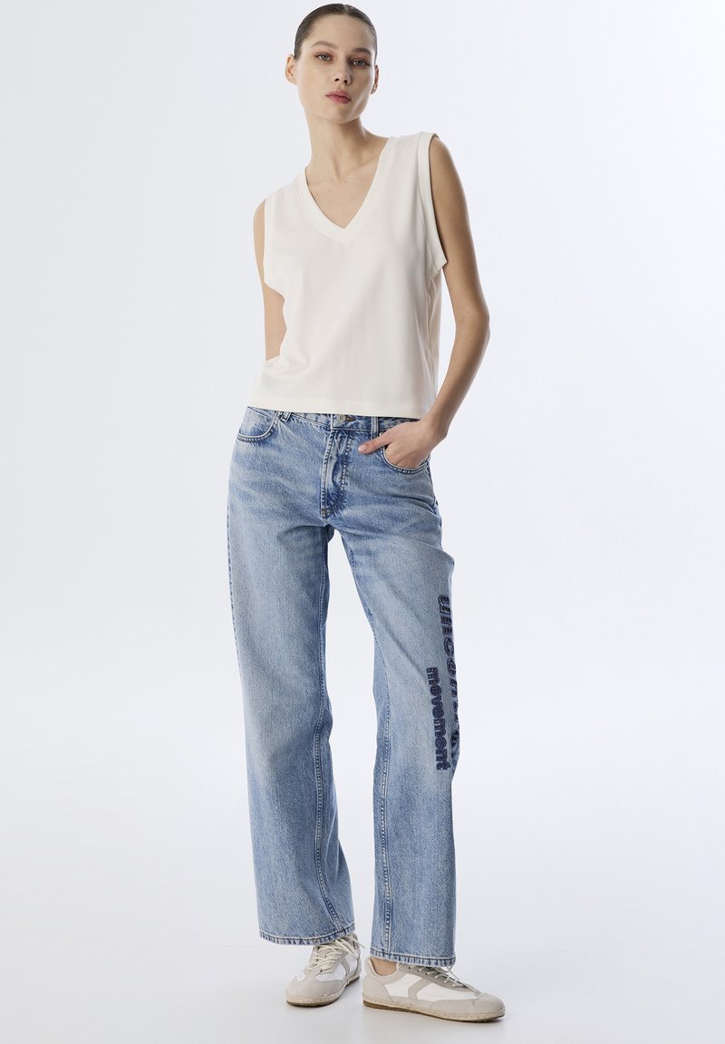 Top bianco senza maniche con scollatura a V, abbinato a jeans dritti azzurri chiari con testo blu scuro sul lato sinistro. Scarpe da ginnastica con texture.