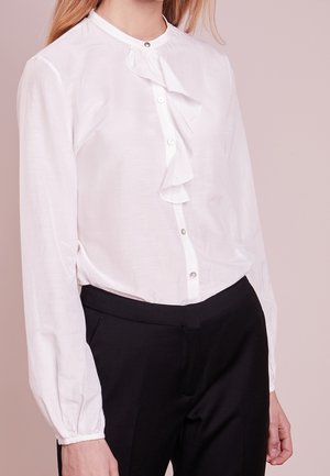 Femme portant un chemisier blanc à manches longues avec des détails à volants et un pantalon noir sur un fond beige uni.