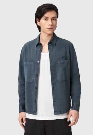 Ung mand iført en mørkeblå skjorte med knapper over en hvid ribbet T-shirt, stående mod en ensartet lys baggrund.