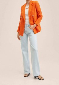 Žena v jasně oranžovém dvouřadém saku, bílém triku, světlých modrých širokých džínách a černých sandálech na podpatku s otevřenou špičkou, stojící s jednou rukou v kapse.