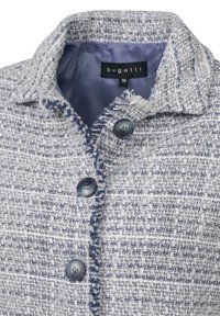 Textured, woven jacket in white and navy with a collar, featuring two blue buttons and a frayed edge along the opening. Lined in light blue.