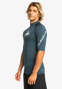 Short-sleeve rash guard in dark teal, made of stretchy material. Features a white logo on the chest and branding along the right sleeve.
