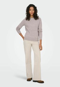 Un pull en tricot gris clair avec un col rond, associé à un pantalon large de couleur crème et des chaussures marron. Le modèle se tient avec une main sur la hanche.