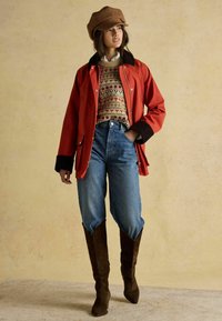 Punane jakk musta kraega, mustriline kampsun, kõrge vöökohaga sinised teksad ning pruunid nahast põlveni saapad. Pruun nokamüts peas.