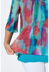 Top colorato e leggero con un motivo astratto trasparente in stile acquerello nei toni del rosso, blu e verde, con un sottogiacca solido di colore turchese.
