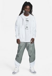 Casaco branco com fecho éclair sobre uma t-shirt gráfica branca com texto preto. Calças desportivas cinzentas e brancas em blocos de cor. Ténis com detalhes claros.