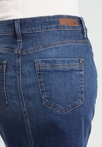 Tmavě modré džíny z denimu s pěti kapsami, viditelným šitím a hnědou koženou značkovou nášivkou na zadním pasu.