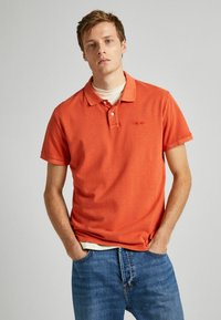 Oranžové polo tričko s krátkým rukávem vyrobené z bavlny, se dvěma knoflíky na zapínání, decentní výšivkou loga a volným střihem. Nosené s modrými džíny.