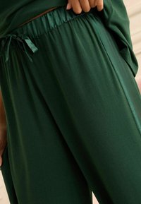 Pantalones deportivos verdes con una textura sedosa, que cuentan con una cintura fruncida y un detalle decorativo de lazo. La tela tiene un acabado liso y elegante.