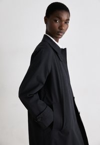 Abrigo negro tipo trench con un cuello clásico, bolsillos laterales y puños abotonados. La tela parece suave y ligera.