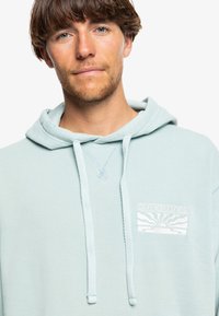 Light blue hooded sweatshirt featuring a front pocket, drawstrings, and a white embroidered Quiksilver logo with sun and wave design.
