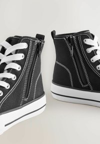 Zwarte canvas hoge sneakers met witte veters, zwarte ritsen en witte stiksel-details. Heeft een dikke witte zool met zwarte accenten.
