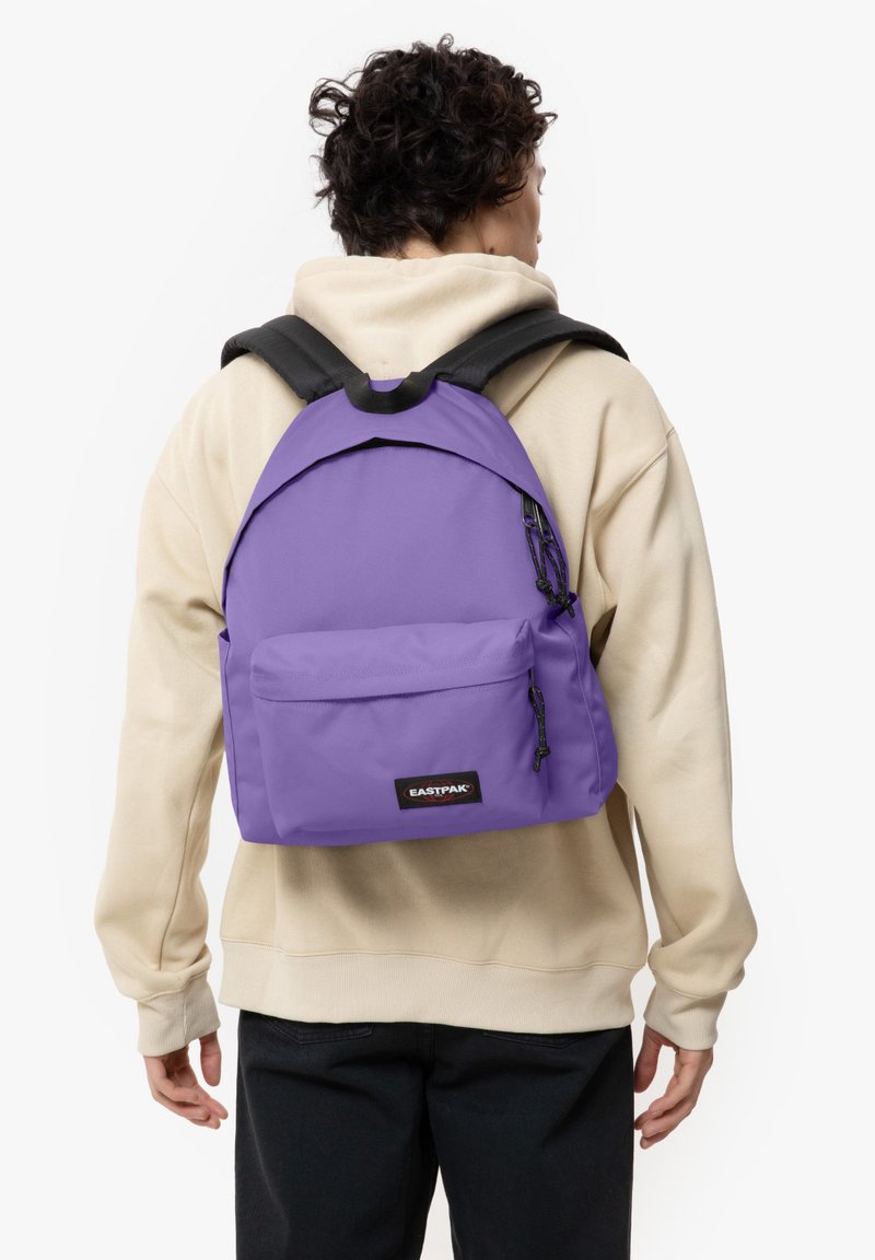 Mochila púrpura con una textura suave, bolsillo delantero con cremallera, correas ajustables y un parche con el logotipo. El material es duradero y está diseñado para el uso diario.