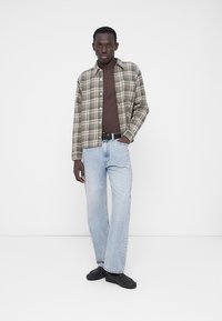 Camicia di flanella a quadri in toni terra, indossata sopra una maglia marrone, abbinata a jeans a gamba dritta azzurri chiaro e sneakers nere.