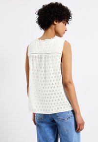 Witte mouwloze top met oogjesborduurwerk, gerimpelde halslijn en gathered achterkant. Gecombineerd met lichtblauwe jeans, met een ontspannen pasvorm.