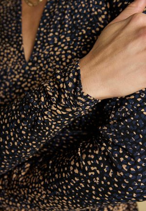 Frau trägt ein langärmliges schwarzes Kleid mit beigen, unregelmäßigen Punktmustern, Hand ruht in der Nähe des Schlüsselbeins, Ausschnitt sichtbar.