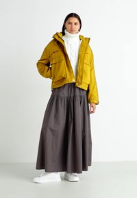 Gele cropped jas, witte kabelgebreide coltrui, lange grijze rok en witte sneakers. De jas heeft voorzakken en een capuchon.