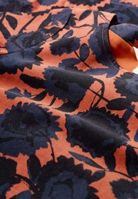 Oranžs un tumši zils ziedu raksts audumā; tam ir mīksta tekstūra, ar lieliem tumši ziliem ziediem un zaļām lapām uz teksturēta fona.