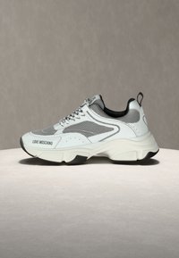 Λευκό και γκρι chunky αθλητικό παπούτσι Love Moschino με διχτυωτά πάνελ, παχιά σόλα και ταινία έλξης σε ουδέτερο φόντο.