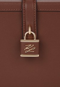 Pruuni nahast käekoti detail, mis sisaldab kullatud lukku koos allkirja logoga. Sujuv tekstuur ja selged jooned iseloomustavad disaini.