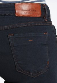 Vaqueros de mezclilla azul oscuro con una etiqueta de cuero marrón en la cinturilla y detalles de costura naranja en los bolsillos.