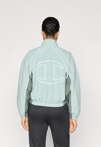 Φωτεινό πράσινο αντιανεμικό μπουφάν με ψηλό γιακά, ελαστική ζώνη στη μέση και μεγάλο λευκό λογότυπο στην πλάτη· διαθέτει αντιθετικές λευκές ραφές.