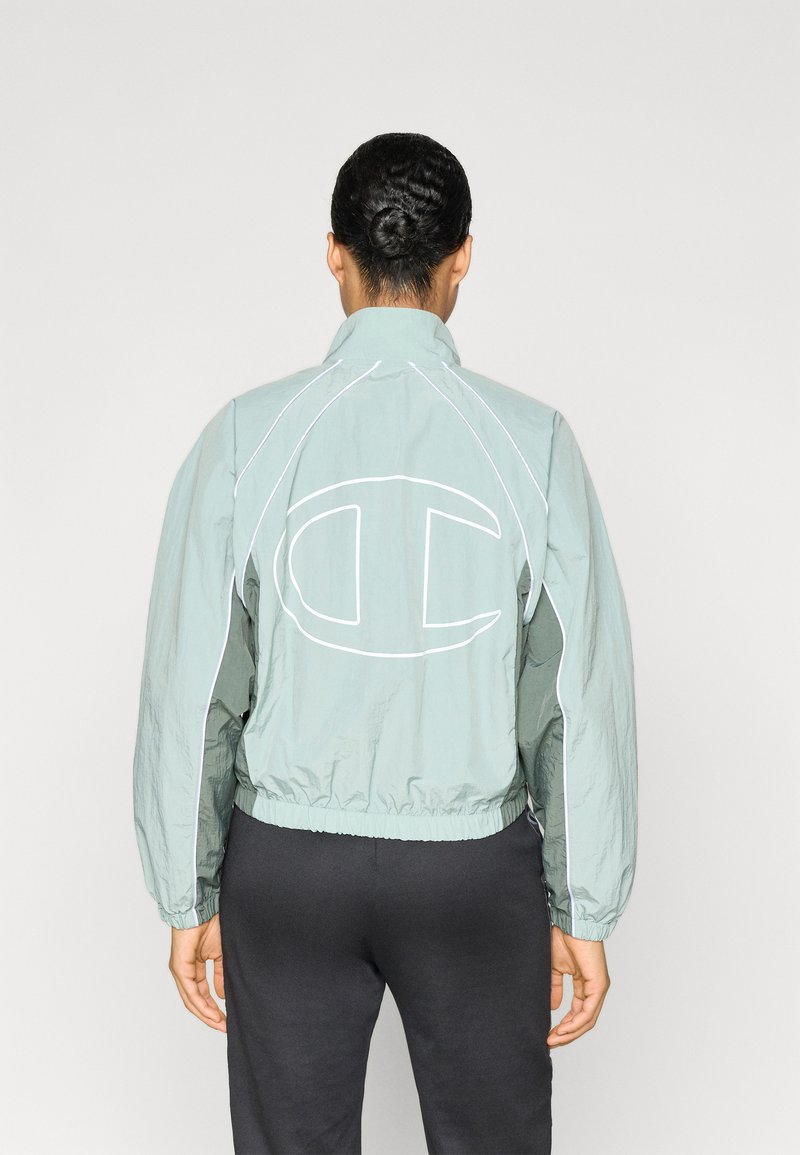 Φωτεινό πράσινο αντιανεμικό μπουφάν με ψηλό γιακά, ελαστική ζώνη στη μέση και μεγάλο λευκό λογότυπο στην πλάτη· διαθέτει αντιθετικές λευκές ραφές.