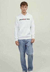 Jovem vestido com um hoodie branco da "Jack & Jones" e jeans azul claro, posando contra um fundo liso de tom bege claro.
