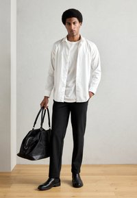 Chemise blanche à boutons superposée sur un t-shirt blanc, pantalon noir fuselé, bottes noires et un sac de sport noir avec poignées.