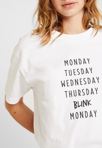 Bílý bavlněný tričko s černým textem, který říká "PONDĚLÍ," "ÚTERÝ," "STŘEDA," "ČTVRTEK," "MRKNUTÍ" a "PONDĚLÍ." Krátké rukávy.