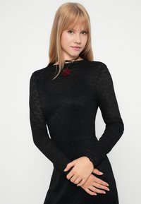 Μαύρο φόρεμα με μακριά μανίκια και υφή λουλουδιών; διαθέτει ψηλό γιακά και ένα pendant από κόκκινο τριαντάφυλλο σε μαύρο κορδόνι γύρω από το λαιμό.