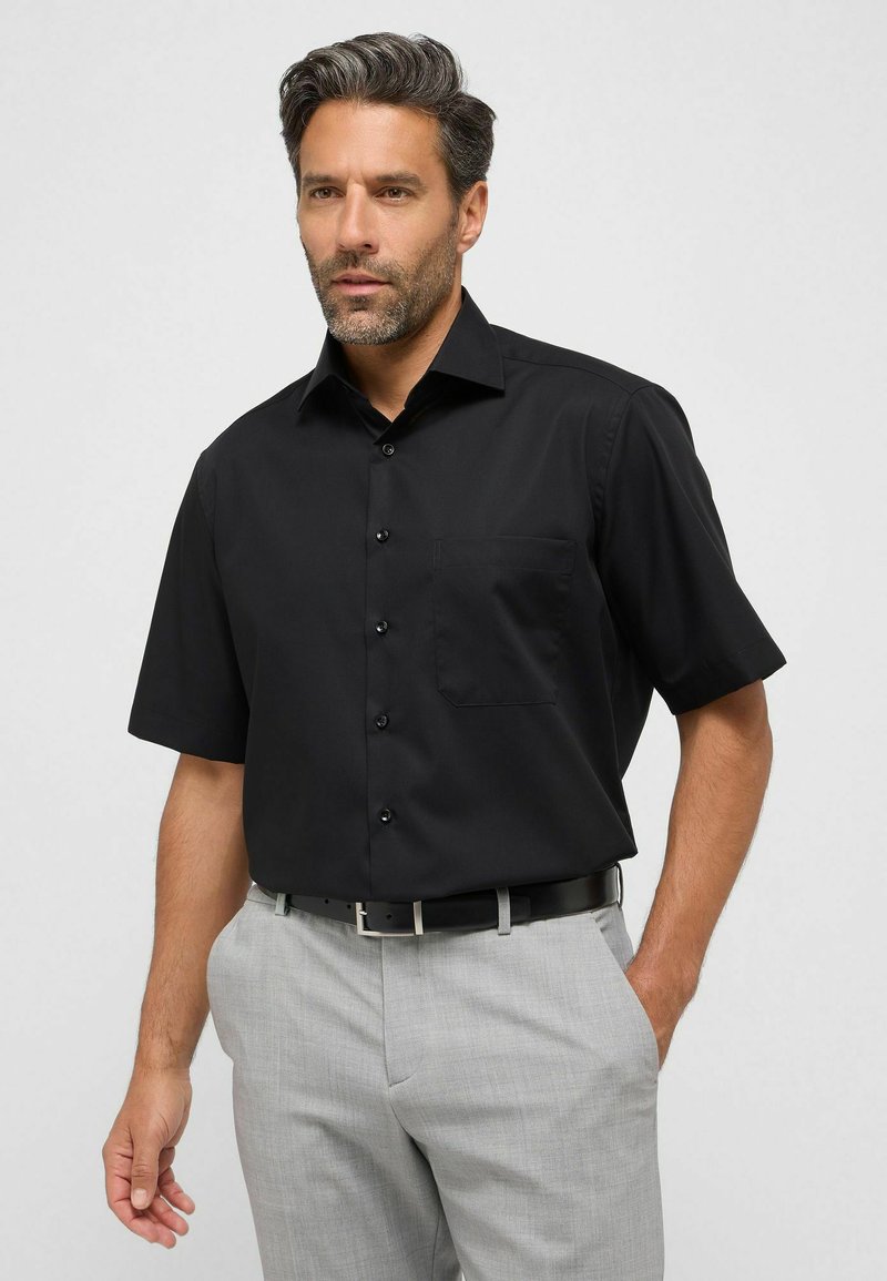 Camicia nera a maniche corte con colletto a punta, dotata di taschino sul petto e tessuto dalla texture liscia, abbinata a pantaloni grigi chiari.