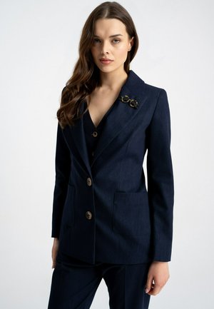 Kobieta o długich brązowych włosach, ubrana w ciemnogranatową marynarkę z broszką w podwójne koło w kolorze żółwia oraz w pasujące spodnie, na białym tle.