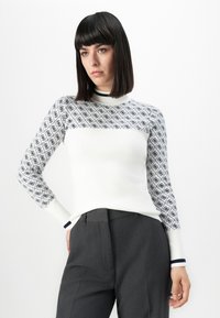 Donna con capelli scuri e lisci, che indossa un maglione bianco con maniche decorate e pantaloni grigio scuro, in piedi con una mano in tasca.
